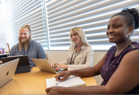 Three people sitting at a table with laptops or notebooks, smiling at and listening to someone off camera