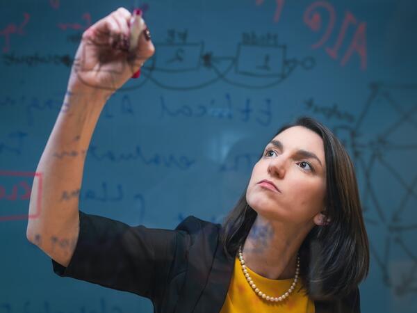 Person in blazer writing on clear board in front of them