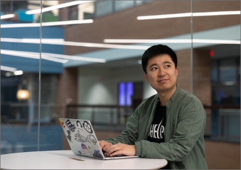 A college student sits in a bright academic building with his laptop.